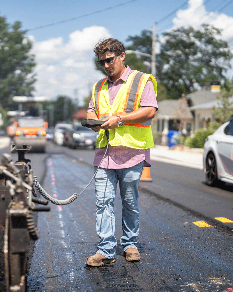 APCC, Employee pink shirt, safety vest, glasses, control, paver, walking on road, 23137_BUILDWITT_2146